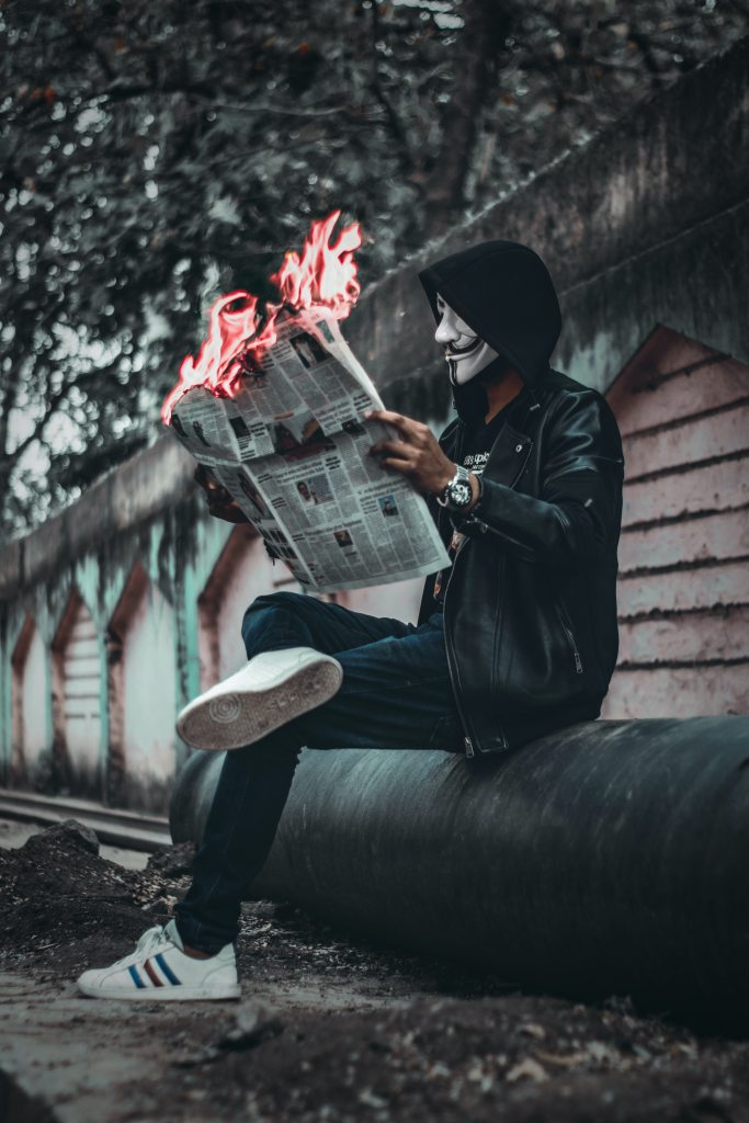 A man in a hoodie and a Guy Fawkes mask, casually reading a newspaper on a park bench, but the newspaper is on fire